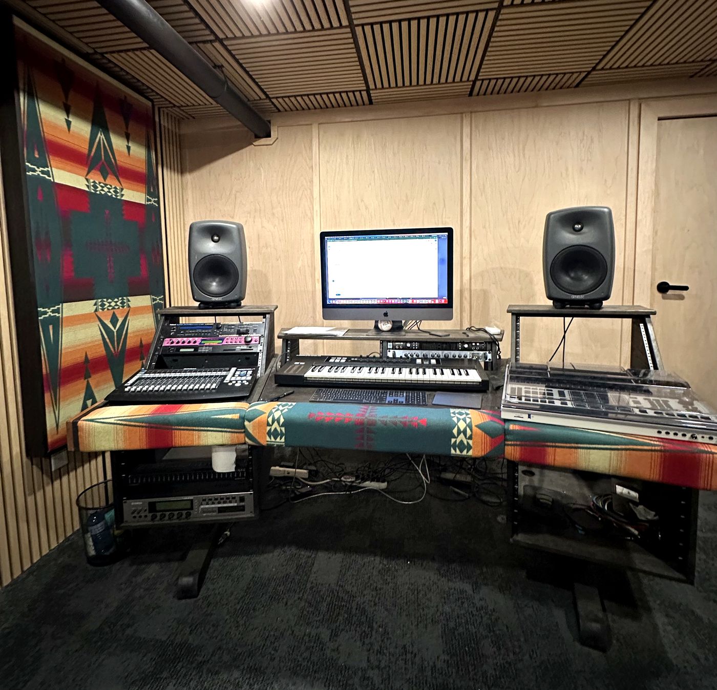 The studio desk face-on: outboard rack units and a digital mixer on the left, a keyboard in the centre and a sampler on the right, with monitors on stands.